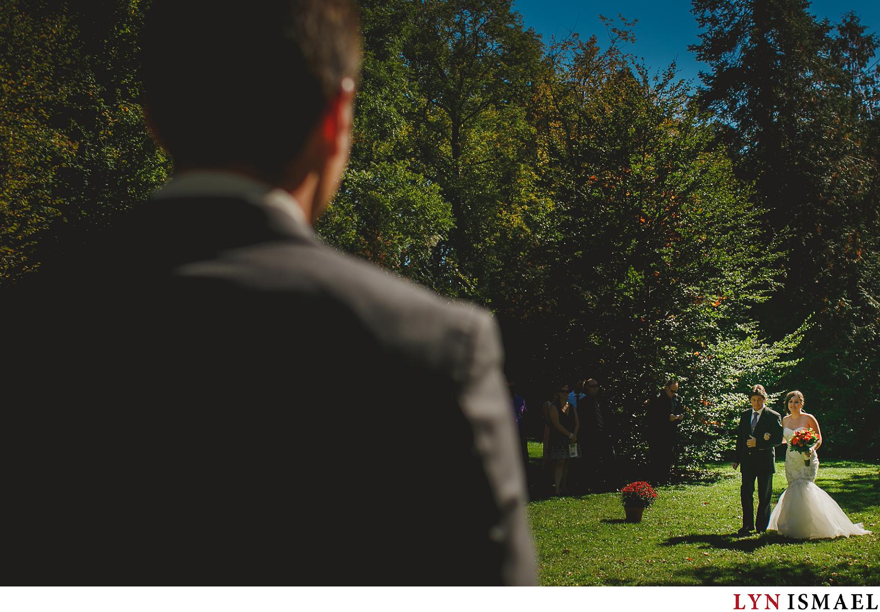 Groom watches his bride walk down the aisle at an outdoor wedding in Elora, Ontario.